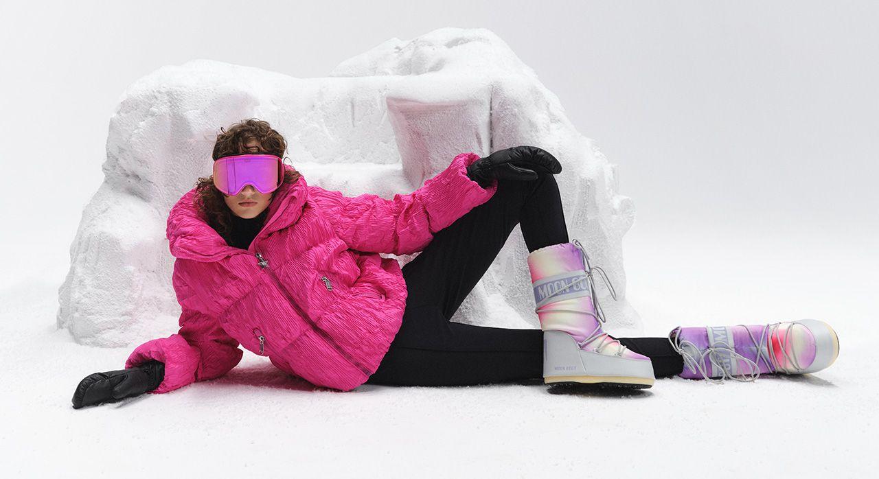 Woman lying on her left side wearing pink ski goggles and jacket and high snow boots